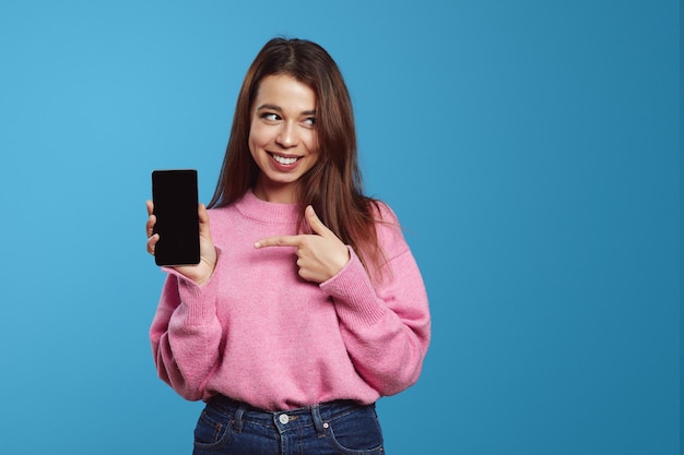 Foto giovane femmina allegra che sorride e distoglie lo sguardo mentre indica lo smartphone con lo schermo vuoto