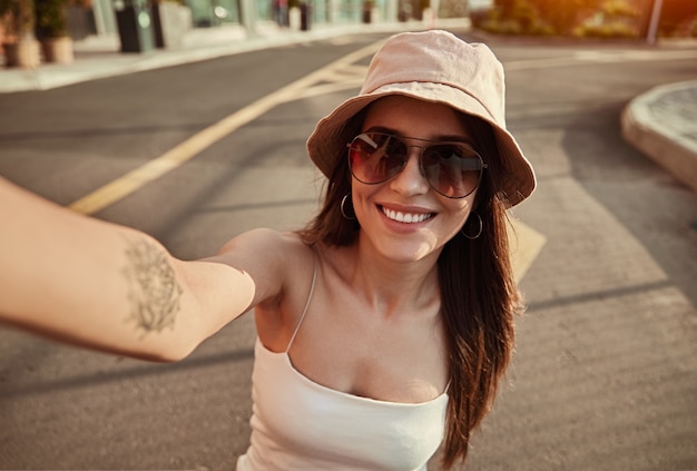 Cheerful young female in stylish sunglasses and hat smiling and taking selfie while enjoying summer day in city