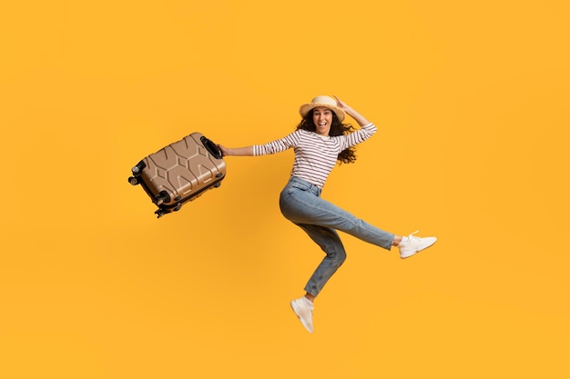 黄色の背景の上の手でスーツケースを持ってジャンプ陽気な若い女性旅行者