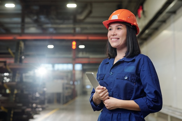 Веселая молодая женщина-инженер с тачпадом стоит в цехе промышленного предприятия, контролируя рабочий процесс