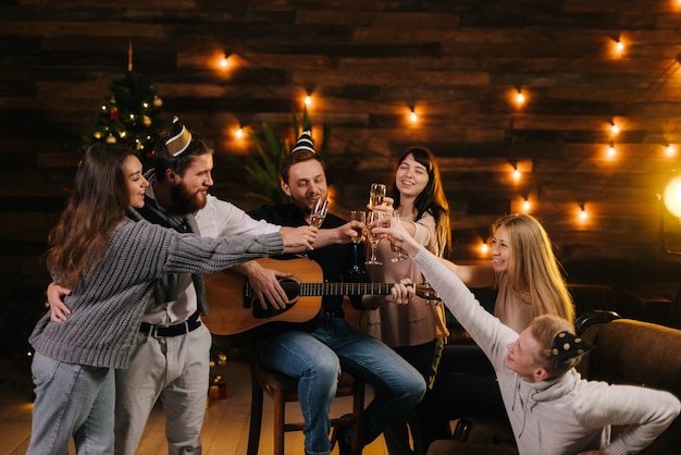 Cheerful young friends are clinking with champagne glasses and celebrating New Year. Christmas tree with garland and festive illumination in background. Friends are celebrating Christmas Eve.