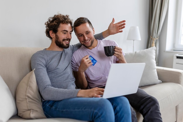 Cheerful young gay couple smiling cheerfully while shopping online at home Two young male lovers using a credit card and a laptop to make a purchase online Young gay couple sitting together indoors