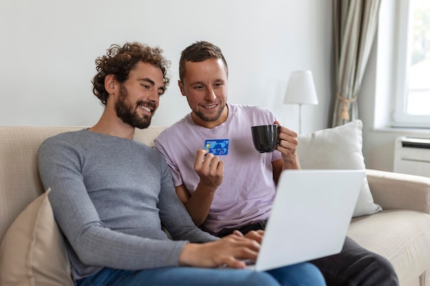 Cheerful young gay couple smiling cheerfully while shopping online at home Two young male lovers using a credit card and a laptop to make a purchase online Young gay couple sitting together indoors