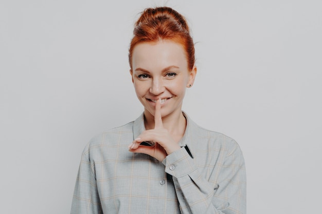 Cheerful young ginger s woman showing hush gesture while posing isolated on grey studio background