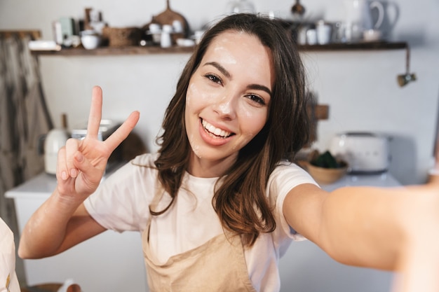 Cheerful young girl covered with flour taking a selfie at the kitchen