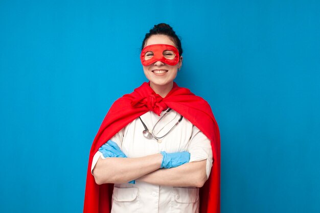 Cheerful young girl doctor in uniform in superman costume on blue background