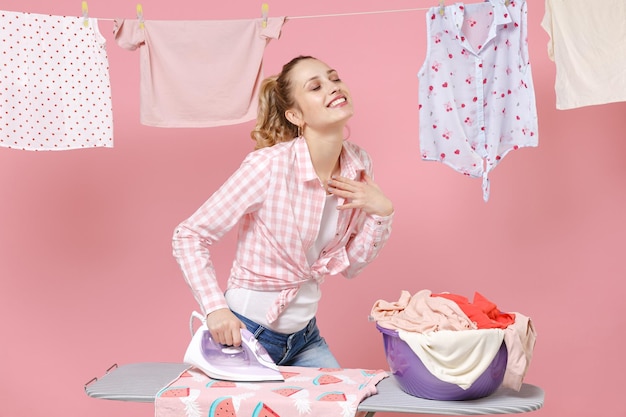 Cheerful young girl housewife in checkered shirt dry clothes on rope ironing clean clothes on board while doing housework isolated on pink background studio. Housekeeping concept. Keeping eyes closed.