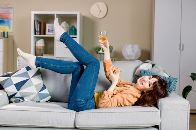 Cheerful young girl in jeans and yellow shirt, holding glass with wine and piece of pizza, while lying on gray sofa in cozy room