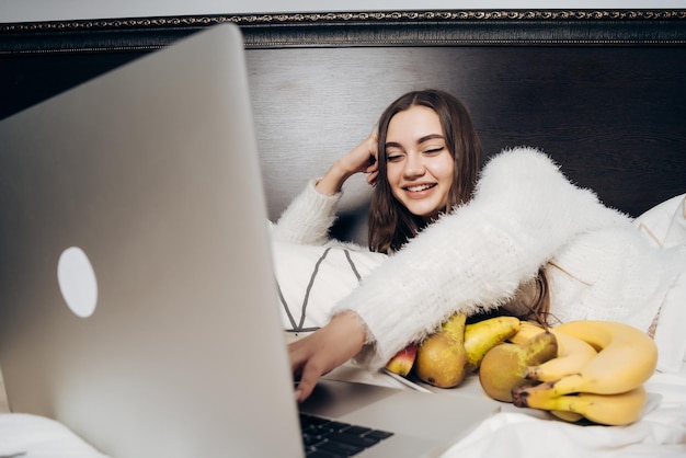 Cheerful young girl lies on the bed relaxes and looks funny ilm on her laptop eats fruit