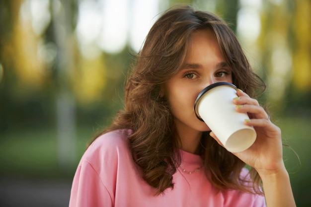 Cheerful young girl in a pink t-shirt walks in the park and drinks takeaway coffee from a paper cup.