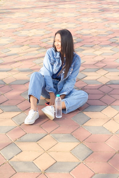 Веселая молодая девушка, сидящая в парке в городе, держит мобильный телефон с бутылкой воды, расположенной на боковой концепции образа жизни