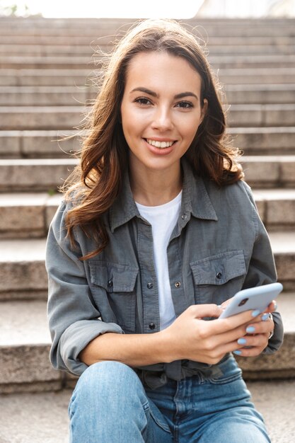 Cheerful young girl sitting on stairs outdoors, using mobile phone