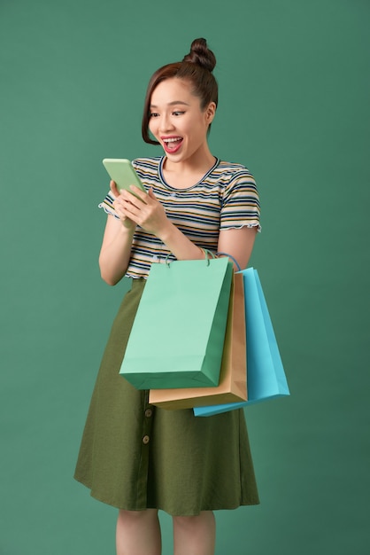 Cheerful young girl standing over green, carrying shopping bags, using mobile phone