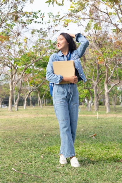 Cheerful young girl standing in the park holding a book and smiling and wears jacket and jeans lifestyle concept