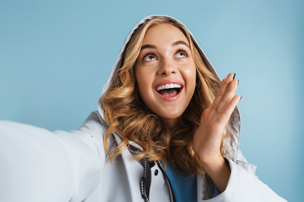 Cheerful young girl wearing raincoat standing isolated, taking a selfie