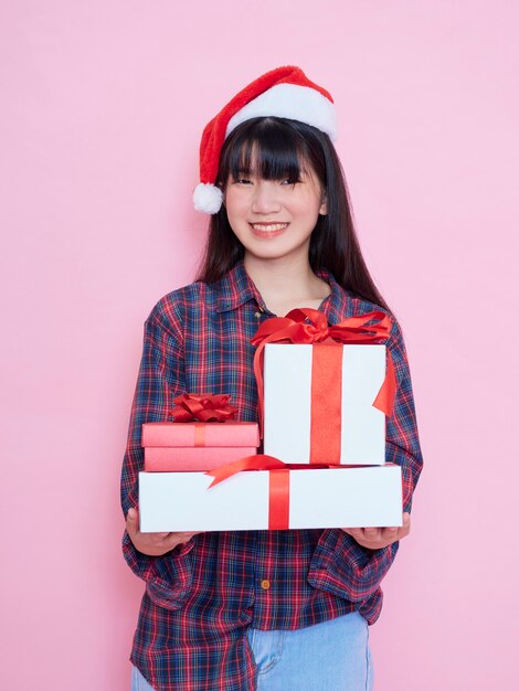 Cheerful young girl wearing santa hat with holding gift boxes on pink background