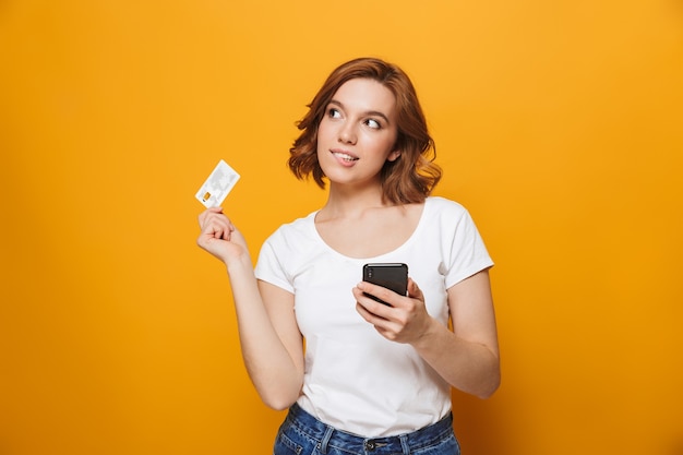 黄色の壁の上に孤立して立っているTシャツを着て、携帯電話を使用して、プラスチックのクレジットカードを示す陽気な少女