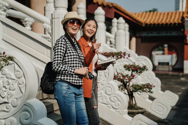 cheerful young girl with guide book in hand pointing away while standing on stairs by marble white stone railings showing friend. two women travelers walking visit in chinese temple in beijing china