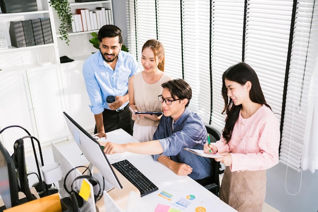 Cheerful young group of Asian businessmen in casual with one sitting and pointing at the computer display and colleagues discussing around him during the meeting Diversity of businessmen in a meeting