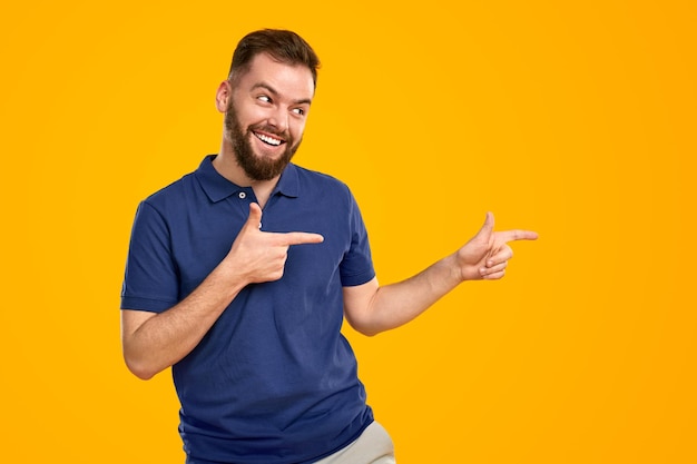 Cheerful young guy in casual outfit pointing at copy space and smiling against yellow background