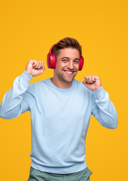 Cheerful young guy clenching fists and smiling while listening to music in wireless headphones and dancing against yellow background
