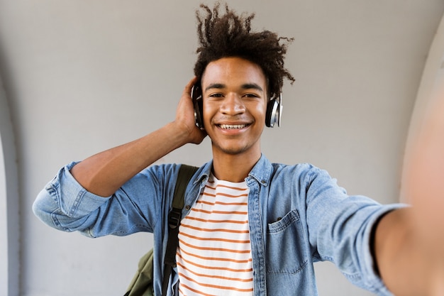 Cheerful young guy standing outdoors listening music with headphones making selfie