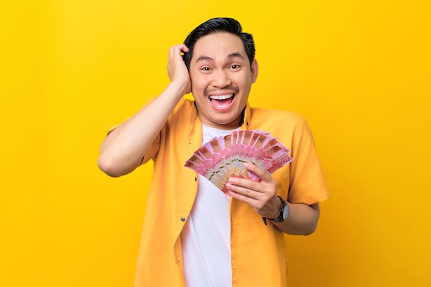 Cheerful young handsome Asian man holding bunch of money banknotes isolated on yellow background