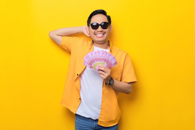 Cheerful young handsome Asian man wearing sunglasses holding fan of money banknotes isolated on yellow background