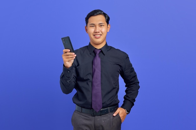 Cheerful young handsome businessman holding smartphone and looking at camera on purple background