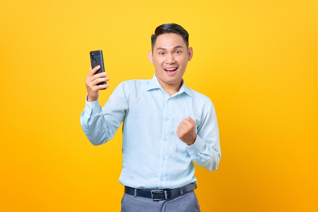 Cheerful young handsome businessman using smartphone and doing victory gesture on yellow background