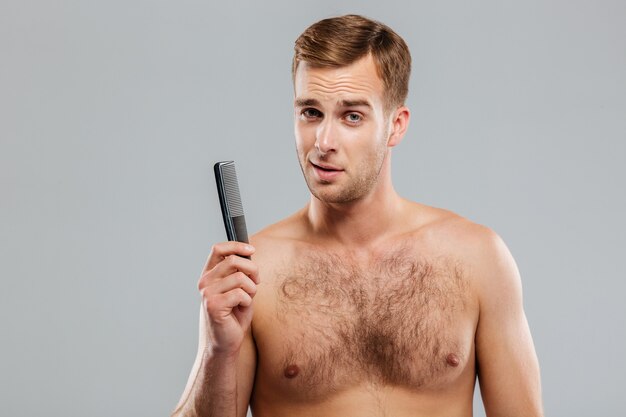 Cheerful young handsome man holding a comb isolated on the gray wall