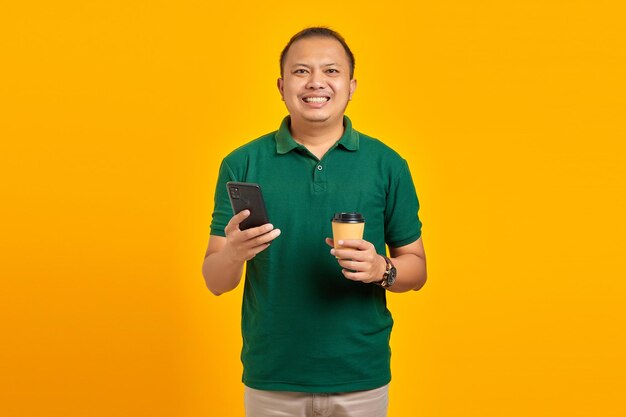 Cheerful young handsome man holding smartphone and cup of coffee on yellow background