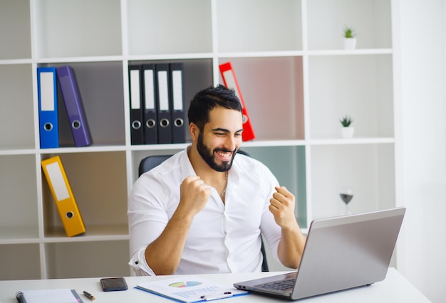 Cheerful young handsome modern director working on a laptop in his large bright office.