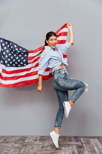 Веселая молодая счастливая девушка держит флаг США и прыгает через серую стену