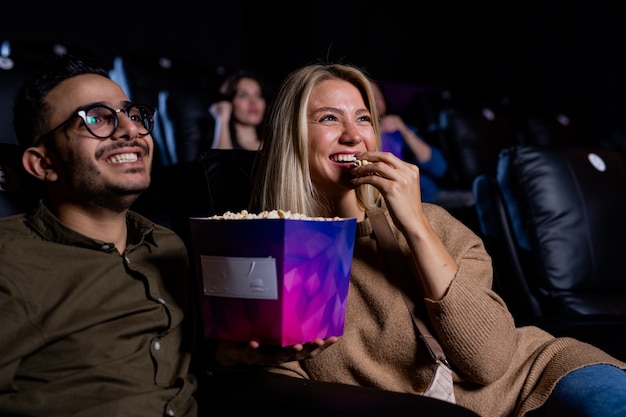 Cheerful young heterosexual couple eating popcorn while watching funny comedy in cinema at leisure