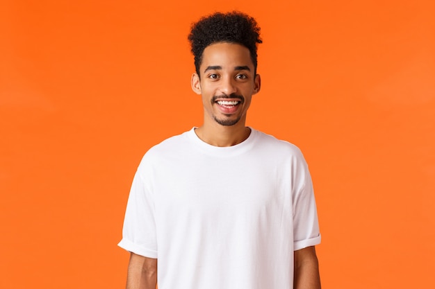 Cheerful young hipster african american guy with moustache and afro haircut, wearing white t-shirt, smiling cute looking  happy, standing orange wall express positivity