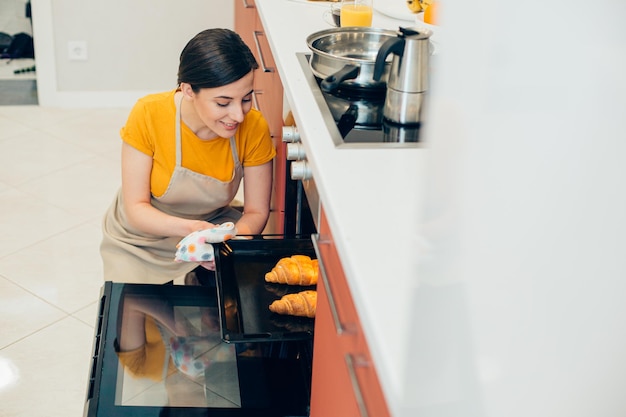 炊飯器のそばのキッチンに座って、パンの上の熱いパン屋を見てエプロンで陽気な若い女性