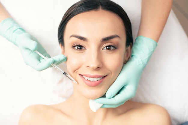 Cheerful young lady feeling good and smiling while getting the injection of hyaluronic acid