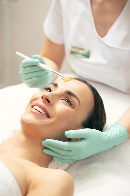 Cheerful young lady having soft brush on her forehead while undergoing facial peeling procedure