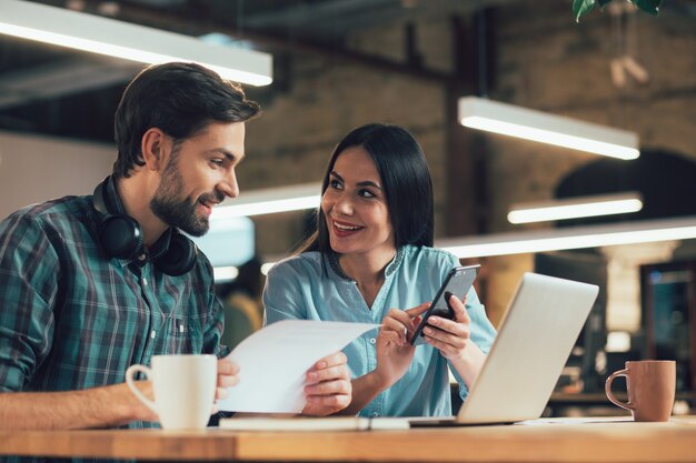 Cheerful young lady sitting at the table next to a gladsome man and showing him something on the screen of her smartphone