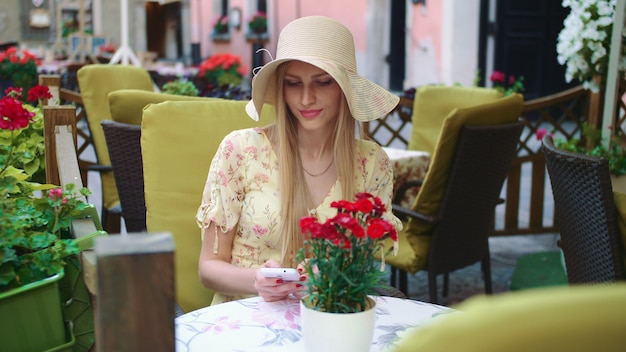 Cheerful young lady sitting at table in outside restaurant and browsing smartphone.