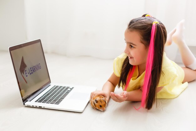 Allegra giovane bambina con un criceto domestico utilizzando il computer portatile che studia attraverso il sistema di e-learning online a casa. formazione a distanza o a distanza