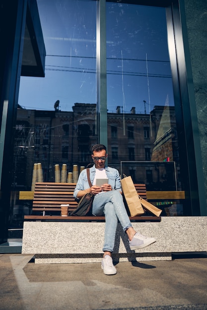 Cheerful young male crossing legs while sitting on the bench and reading new online