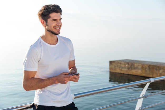 海の近くに立っている携帯電話を使用して白いTシャツを着た陽気な青年アスリート