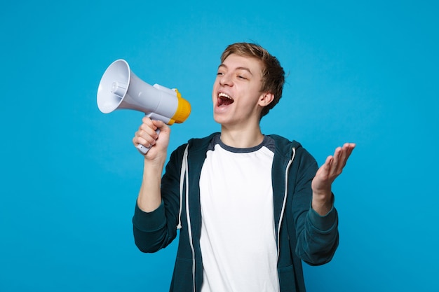 Cheerful young man in casual clothes looking aside, screaming on megaphone, spreading hands isolated on blue wall . People sincere emotions, lifestyle concept. 