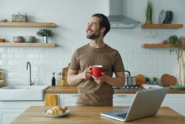 キッチンで時間を過ごす間コーヒーを楽しみ目をそらしている陽気な若い男