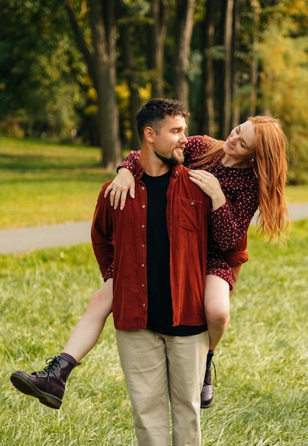 Cheerful young man gives a piggyback ride to his red head woman in a park