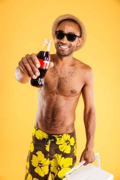 Cheerful young man in hat and sunglasses holding coke bottle and cooler bag over orange wall