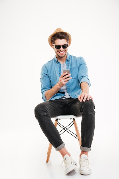 Веселый молодой человек в шляпе и солнцезащитных очках сидит и использует смартфон на белом фоне