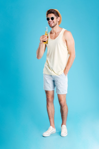 Cheerful young man in hat and sunglasses standing and drinking beer over blue wall